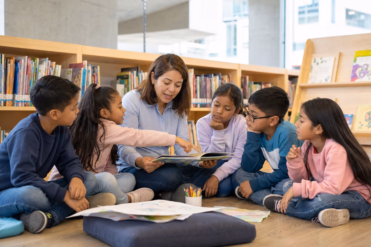 Docente acompaña a un grupo diverso de niñas y niños durante una actividad de lectura en una biblioteca escolar